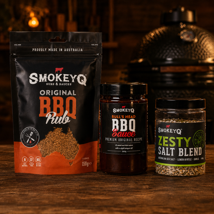 SmokeyQ Original BBQ Bundle met Original BBQ Rub, Bull's Head BBQ Sauce en SPG Salt Blend op houten tafel bij kamado BBQ